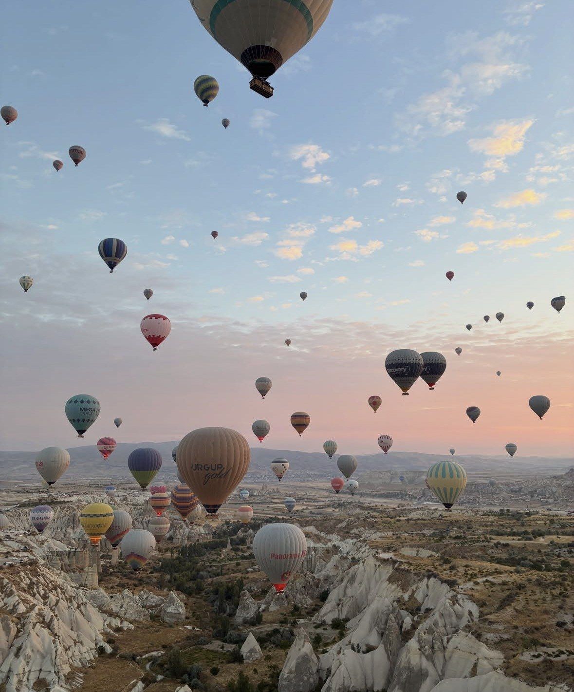 Dense cluster of balloons over Cappadocia cliffs under pink dawn clouds