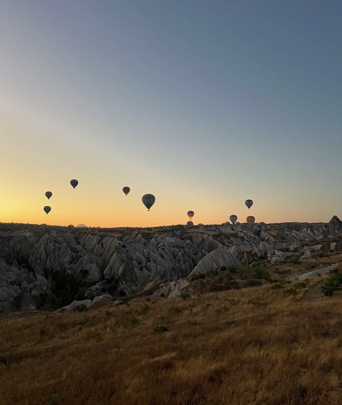 Dry grassy ridge above eroded Cappadocia valley with balloons at sunrise