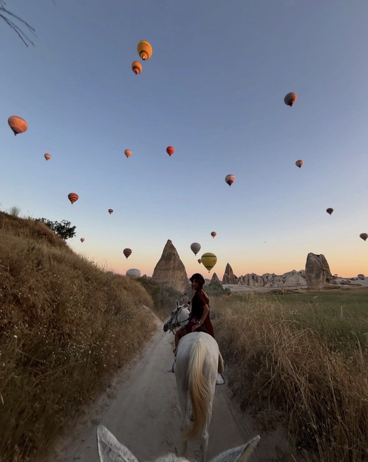 Rider on white horse on narrow Cappadocia trail beneath morning balloons